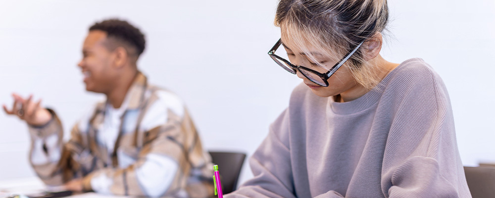 A girl with glasses doing classwork while a guy is chatting in the background