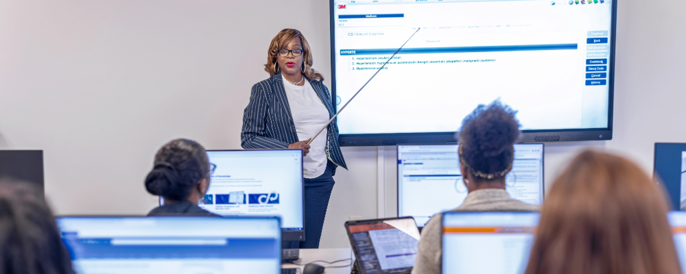 An instructor with a stick pointing to the screen is teaching a classroom full of students